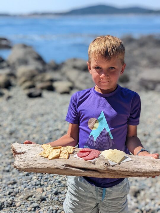 a boy holds a piece of driftwood with meats, cheese, and crackers on top.