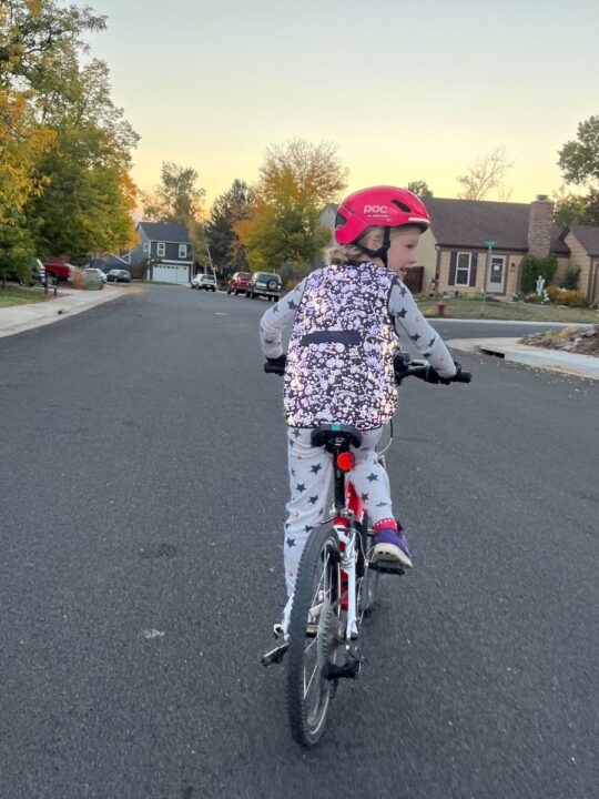 A girl rides a bike down a neighborhood street in star PJs and a reflective vest. 