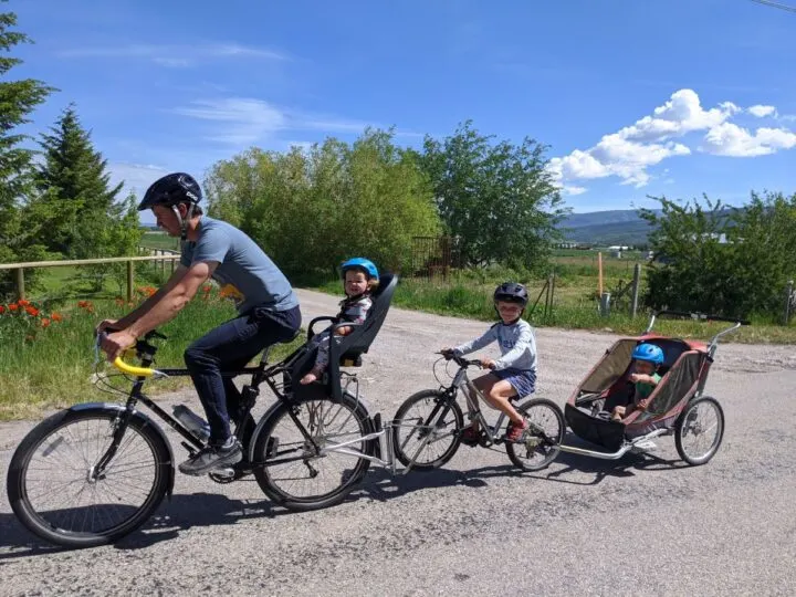 A dad rides his bike with a Burley baby seat on the back, followed by a kids bike hooked on with the follow me tandem, and last, pulling a chariot baby trailer.