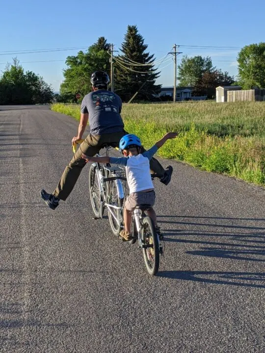 A dad and child ride bikes connected with a follow me tandem, legs and arms flailing in joy.