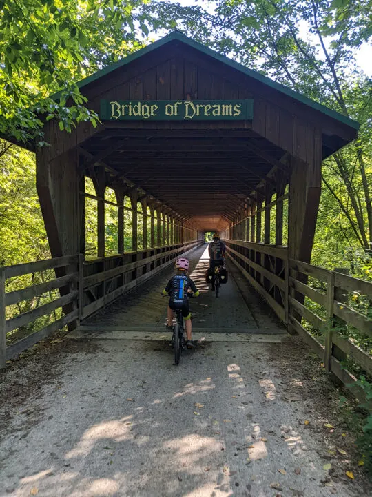 Adult and child ride on bicycles away from photographer across a covered bridge. 
