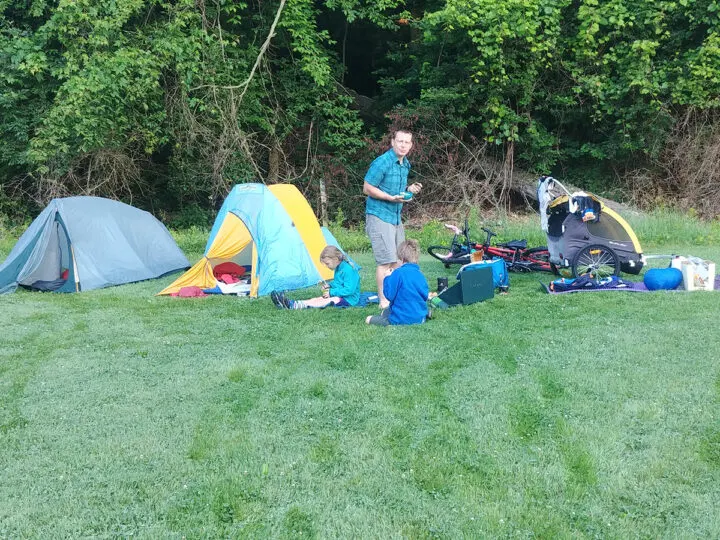 Two tents, a bike trailer, and a lot of gear in the grass with two children sitting and one adult standing.