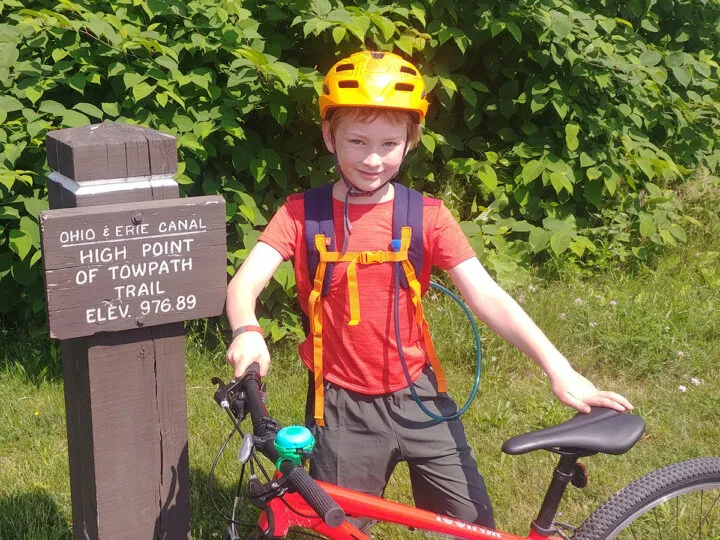 Smiling boy wearing bike helmet and holding a bike standing by a sign that says, "Ohio & Erie Canal High point of the towpath trail elev. 976.89".