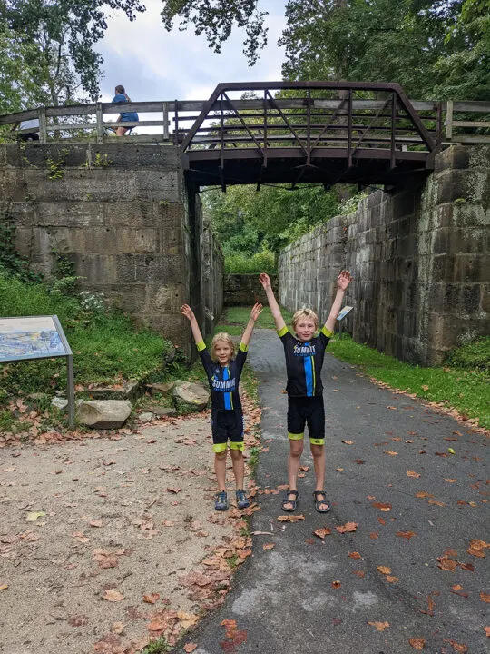 Two kids in cycling clothes with arms in the air in front of a drained canal lock.