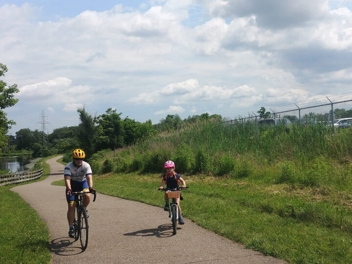 Man and girl ride bikes side by side on a gravel trail winding through a field next to a canal. 