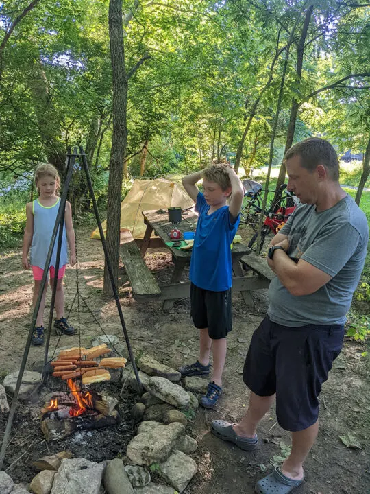 A boy and girl and their father standing by a fire watching hot dogs and buns cook.