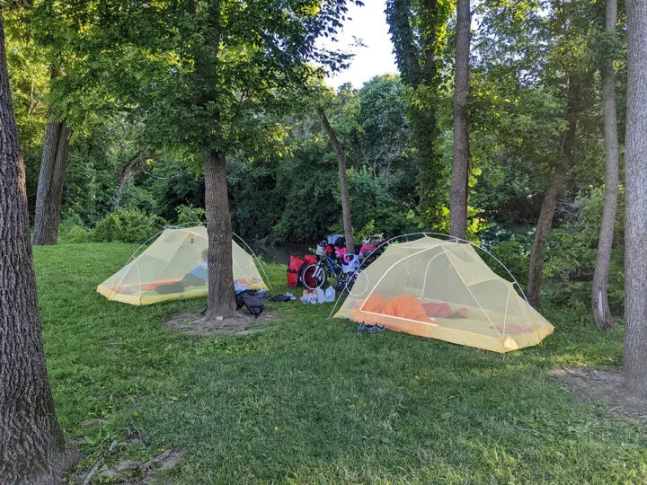 Two mesh tents with one child in each and bicycles and gear between them. The tents are in a stand of trees on grass.