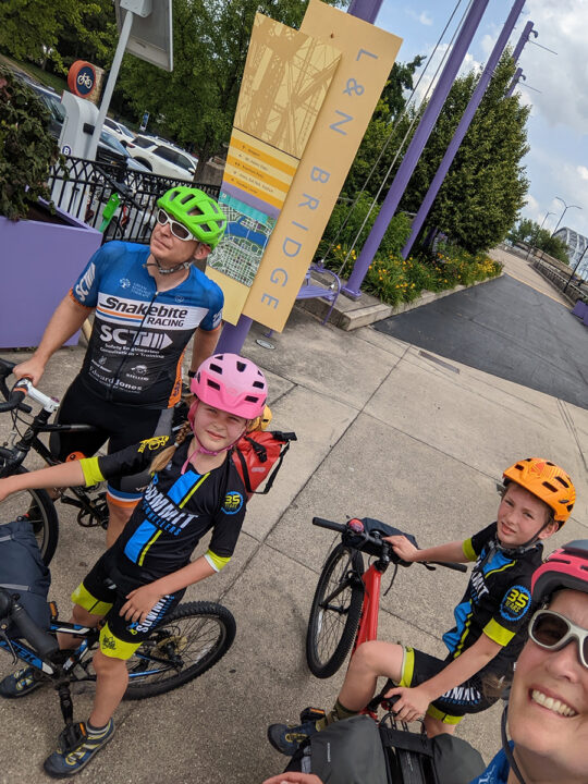 Selfie of a family of four standing over bikes in front of a bridge. 
