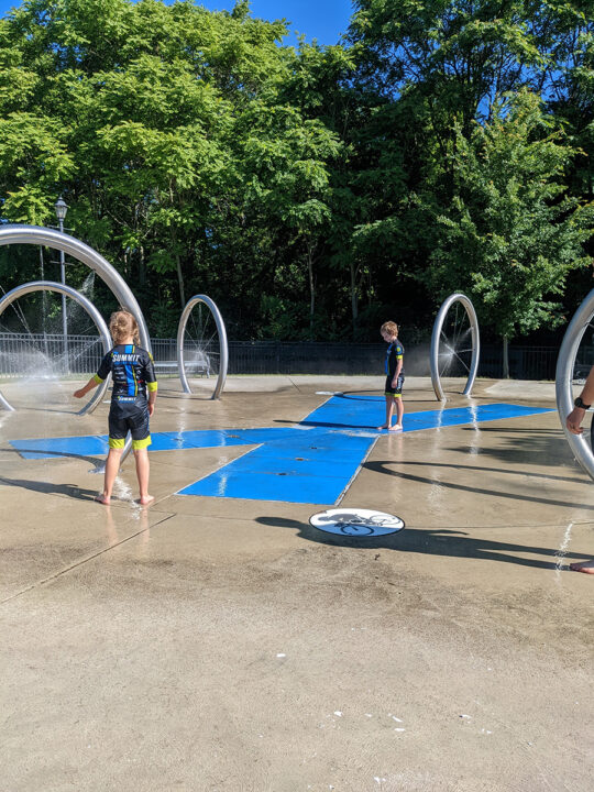 Two barefoot children in cycling clothes play at a splash pad. 
