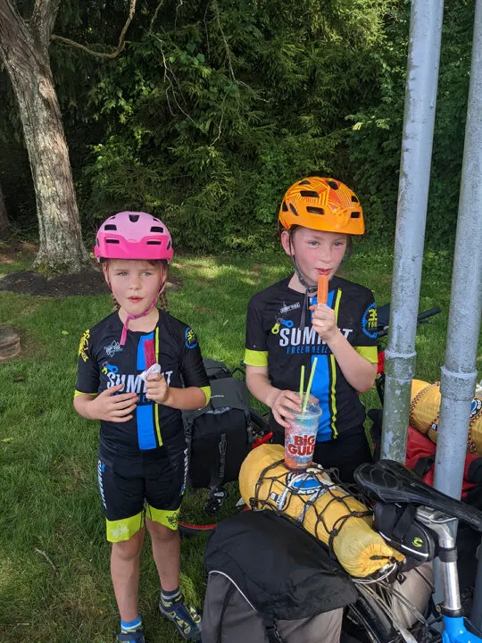 Two children in cycling gear standing near loaded bikes eating popsicles and holding. aBig Gulp cup. 