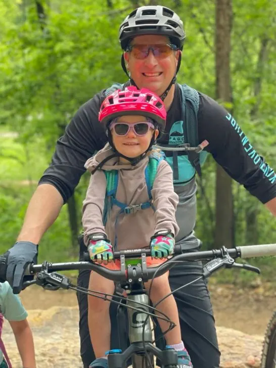 A young girl in a pink helmet sits on a Shotgun mountain bike seat with her original handlebars with her dad behind her. 
