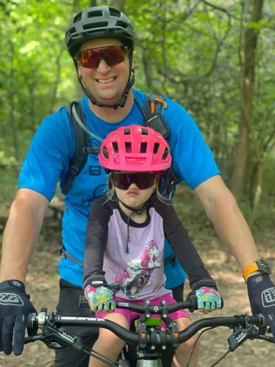 A young girl in a pink helmet sits on a Shotgun mountain bike seat with her pro handlebars with her dad behind her. 