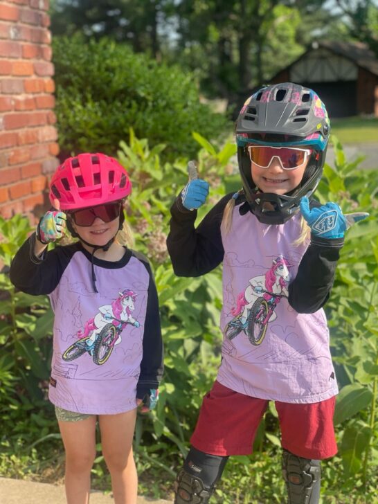 Two young girls wearing shotgun upshift unicorn jerseys are ready for a bike ride.