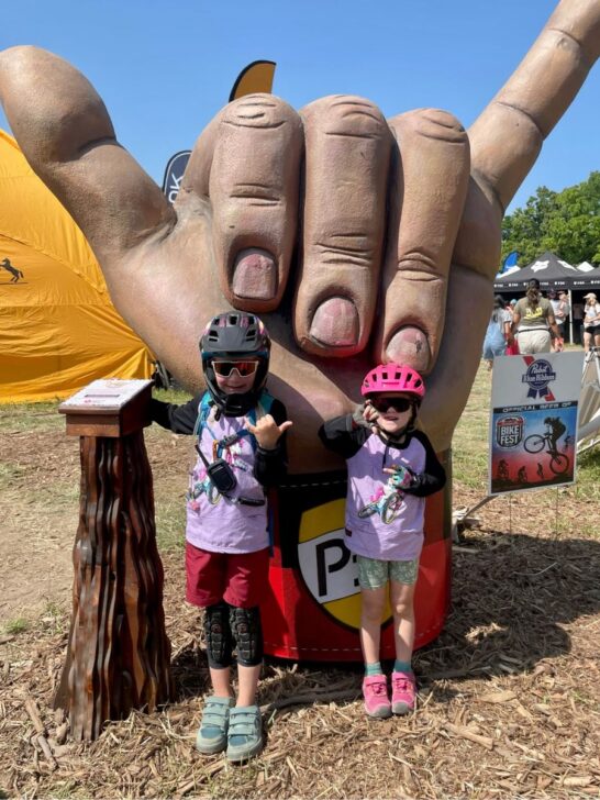 Two young girls wearing a bike helmet and shotgun upshift unicorn jerseys give a shaka sign.