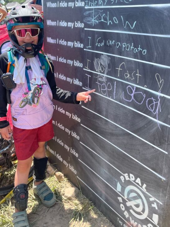 A young girl wearing a bike helmet and shotgun bike jersey points to a chalk board that she wrote on.