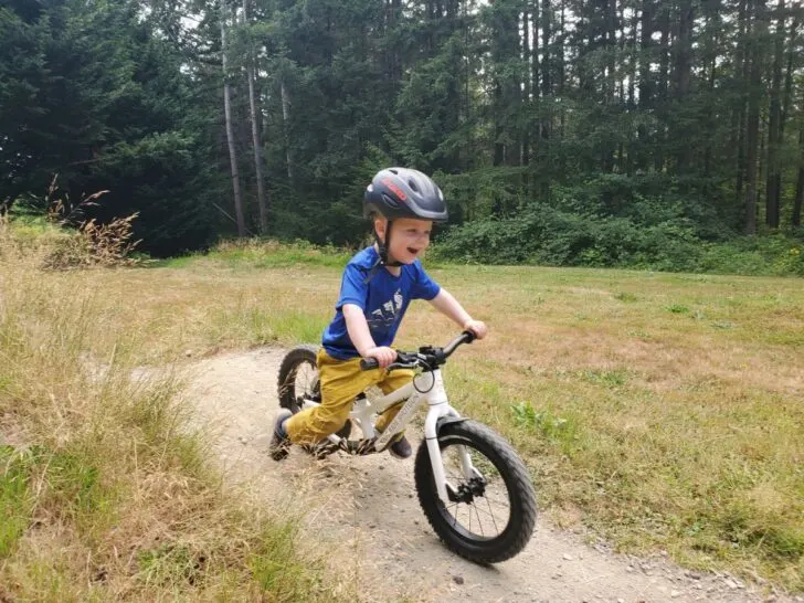 Little boy on a white dirt hero balance bike is coasting around a pump track wear a black helmet and a smile on his face