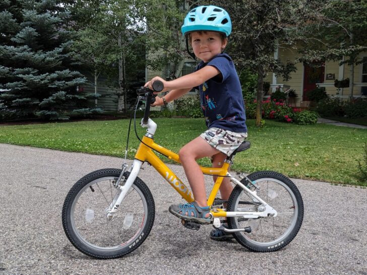 A boy sits on a Woom 3 16 inch yellow bicycle, smiling at the camera.