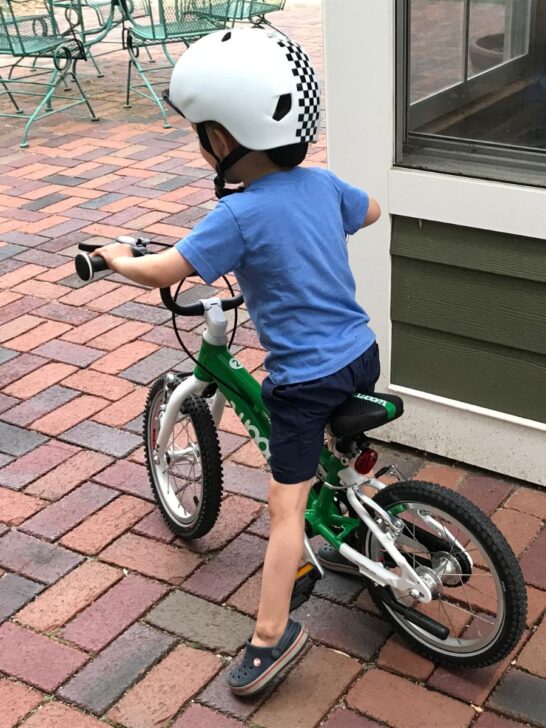 Boy in blue shirt on a green Woom 2 bike on a brick patio.