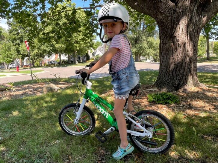 Girl on green Woom bike near a tree.
