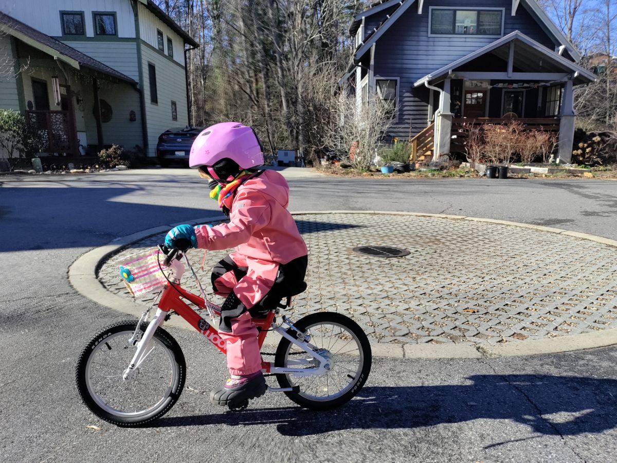 A kid in a pink snow suit and pink helmet rides a red bike in a culdesac