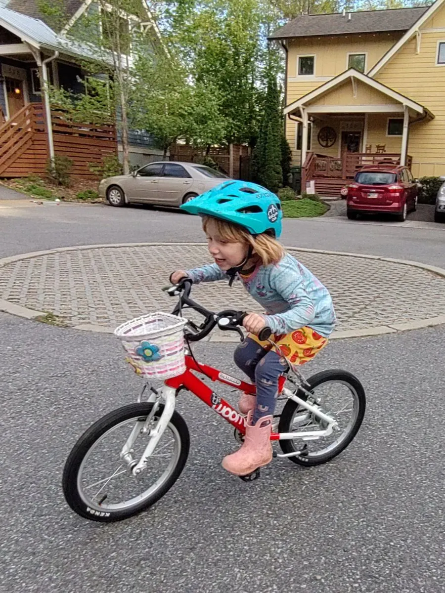A little girl with a blue helmet rides a red bike with a plastic flower basket attached to the front 