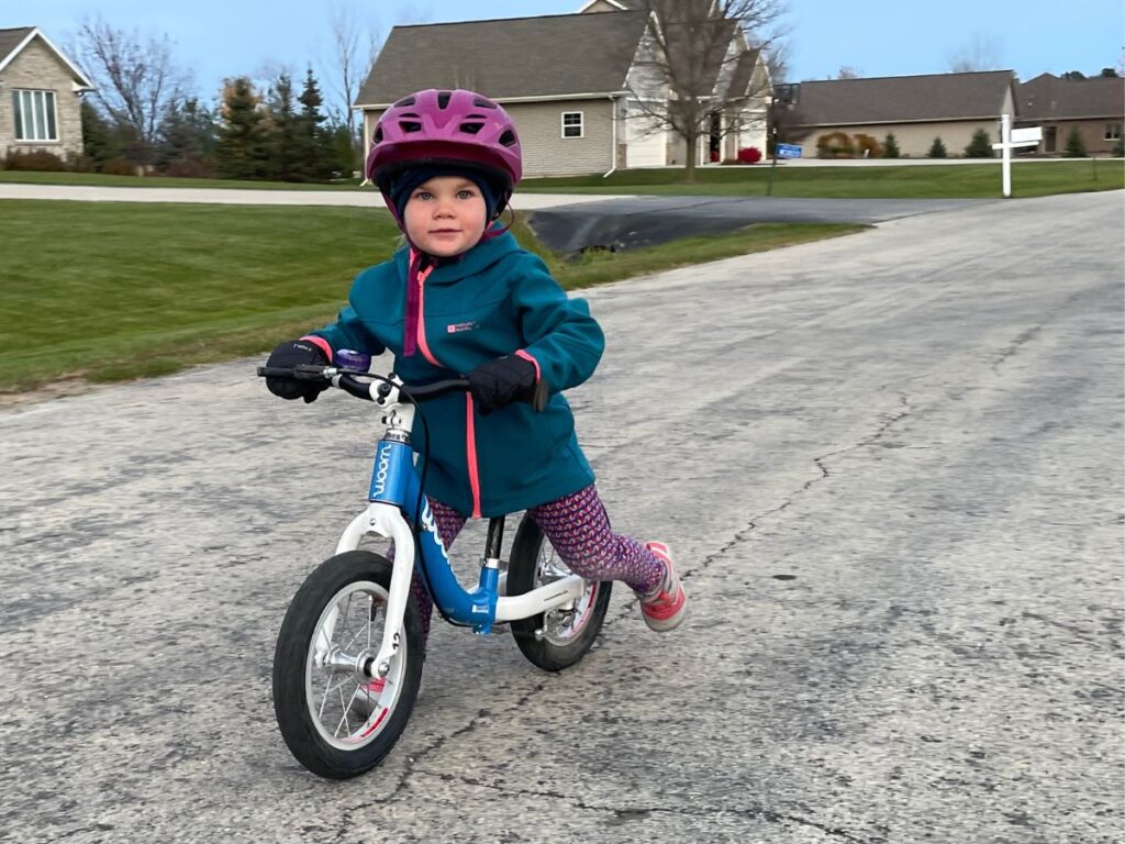 A kid in a blue jacket and pink helmet rides a Blue Woom balance bike