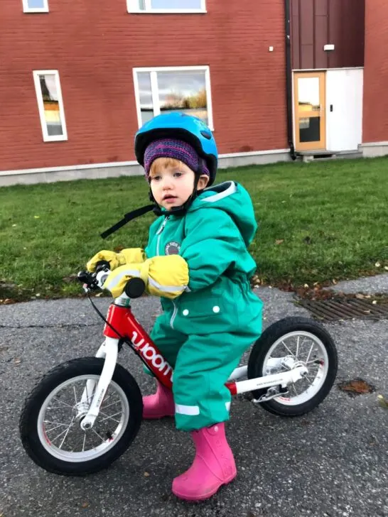 A kid in a green snow suit and yellow gloves with a blue helmet sits on a red Woom balance bike. 