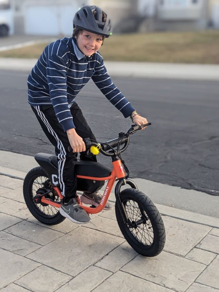 A boy stands on the pegs of an orange Himiway e-bike, smiling for the camera.