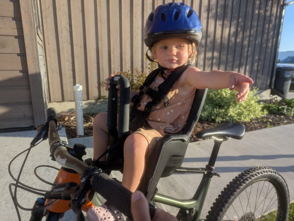 A girl sits in an infant shotgun seat atop a bike.