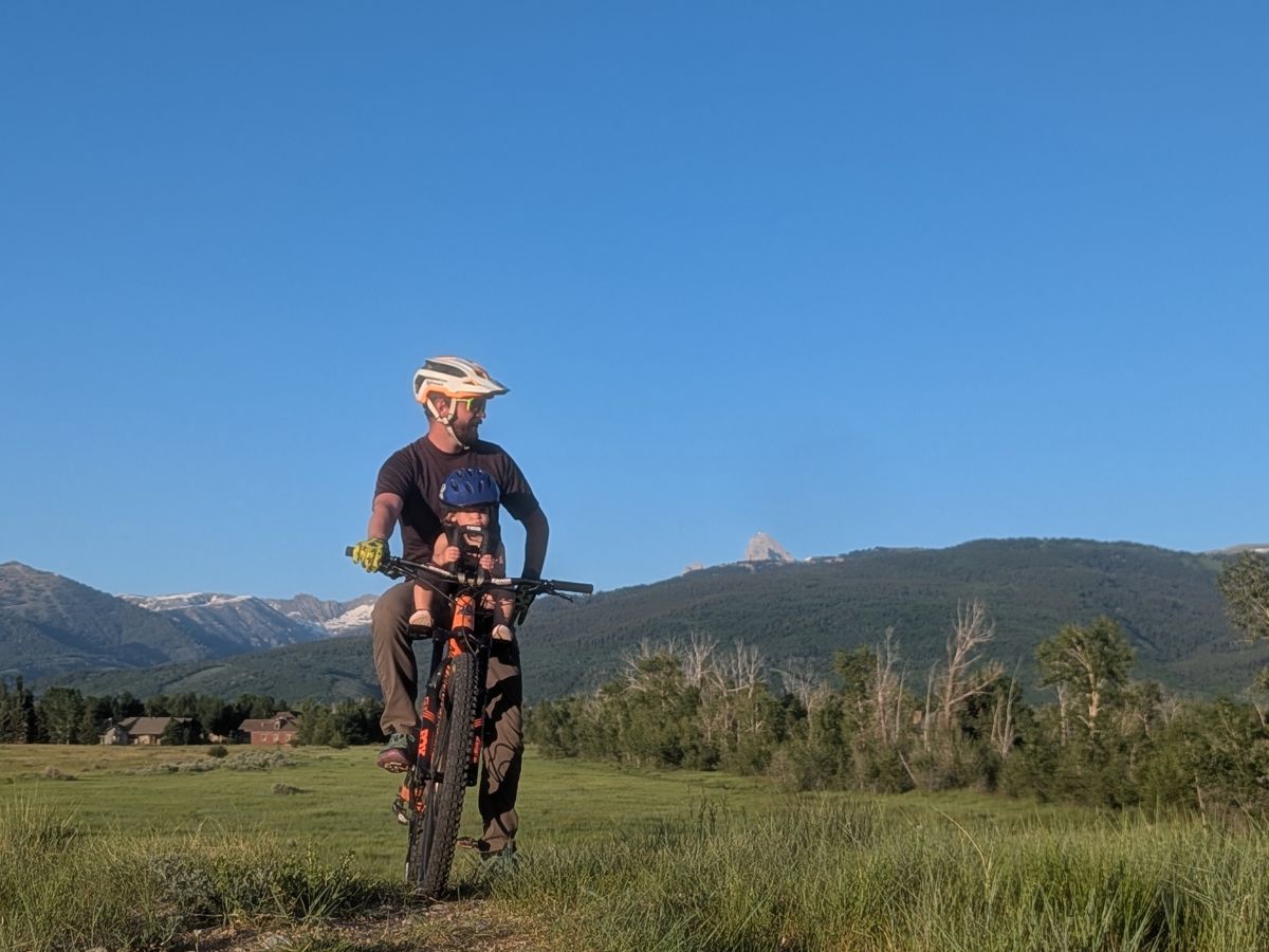 TMM and KRB Horizontal and Featured (31) A dad and baby pose atop a hill on a bike equipped with a front seat. Blue skies, mountains and trees in the back ground.